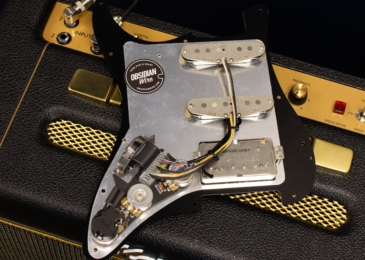 The back side of an HSS loaded Stratocaster pickguard is shown with the wiring harness and pickups. The pickguard is black with aluminium shielding foil, on a black and gold Marshall amp.