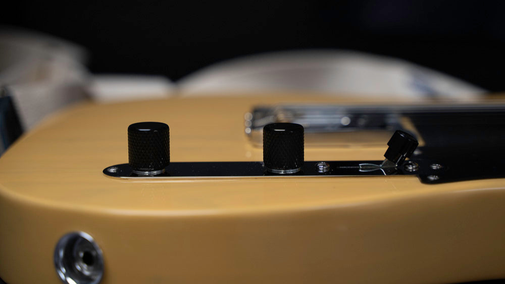 A chrome loaded control plate with black knobs is shown on a Blonde telecaster. The background of the image is blurred