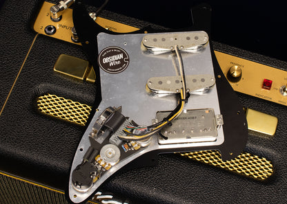 The back side of an HSS loaded Stratocaster pickguard is shown with the wiring harness and pickups. The pickguard is black with aluminium shielding foil, on a black and gold Marshall amp.
