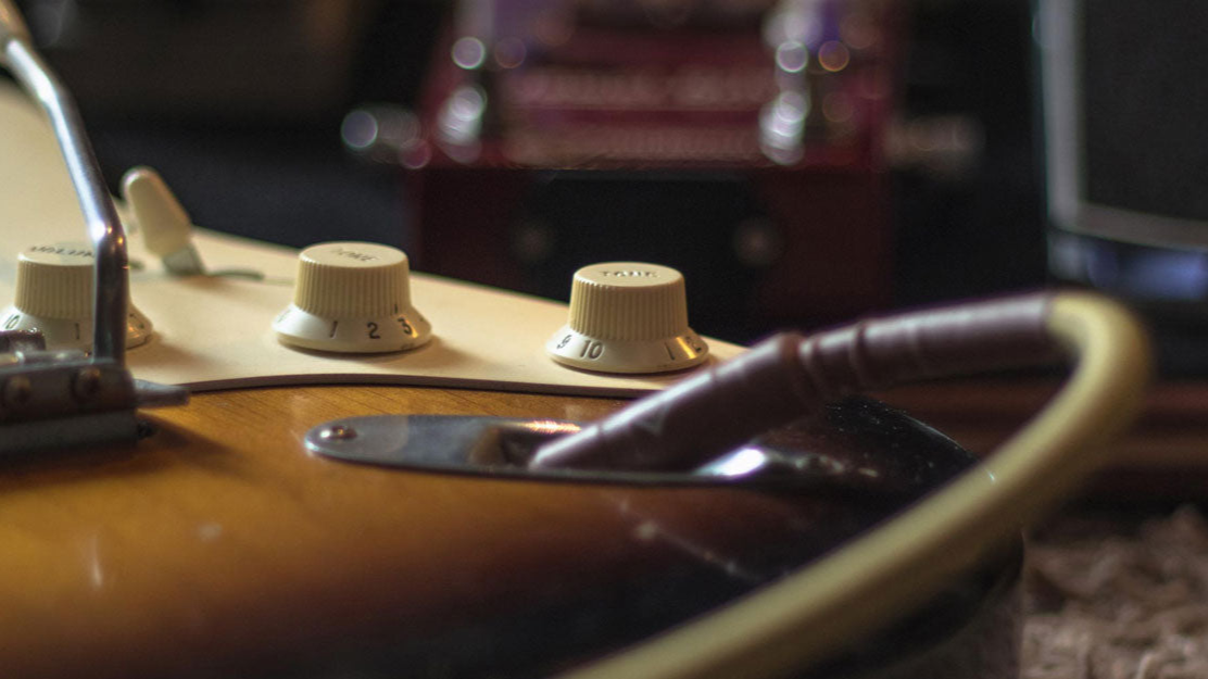 A wide banner image showing the tone and volume controls of a Fender stratocaster. The knobs are aged white, while the body is a traditional three tone sunburst.